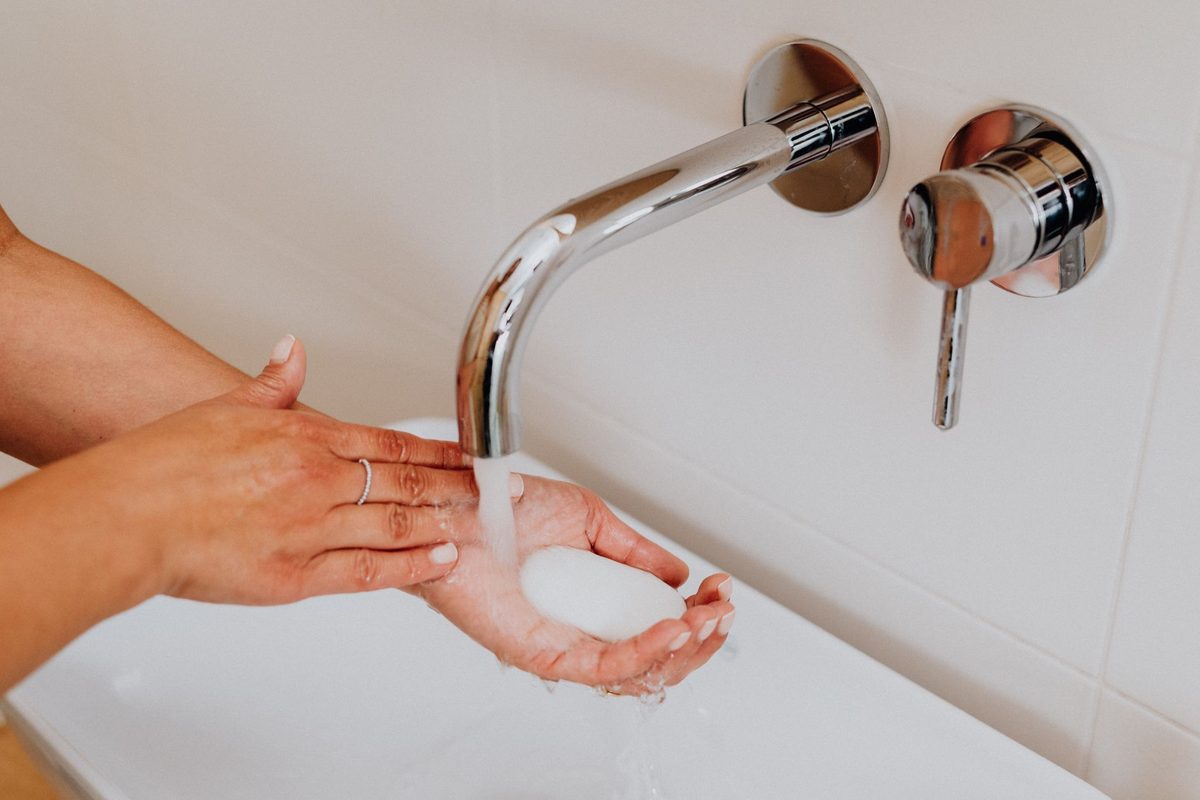 Hands lathering a bar of soap under a running chrome tap while wearing a thin band ring